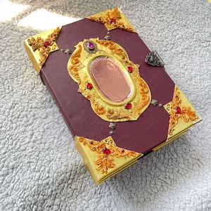 Op de afbeelding: Een bordeauxrood boek met gouden versieringen en rode juwelen. De kaft heeft een groot gouden frame met een ovale spiegel en sierlijke hoekontwerpen. Een decoratieve sluiting sluit het boek af. Afmetingen: 20 cm x 15 cm.