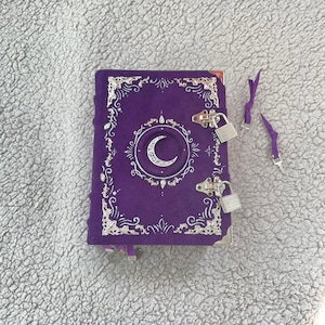 May include: A purple journal with a crescent moon design and silver accents. The journal has a decorative border, corner protectors, and two silver padlocks. Two purple ribbon bookmarks and two keys are also visible.