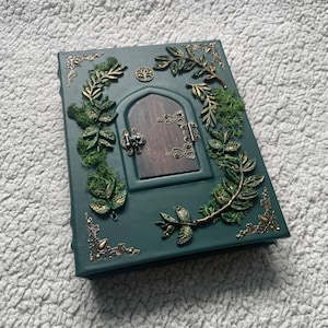 Puede incluir: Una cubierta de libro rectangular de color verde oscuro con un elemento central de puerta arqueada en tono madera oscura. La cubierta está decorada con ramas de hojas doradas metálicas, filigranas en las esquinas, un emblema de árbol y musgo.