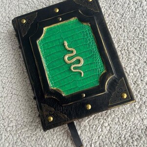 Pode incluir: Um di&aacute;rio encadernado em couro preto com uma capa verde em relevo e um design de cobra dourada. O di&aacute;rio tem um marcador de fita preta e detalhes met&aacute;licos dourados.