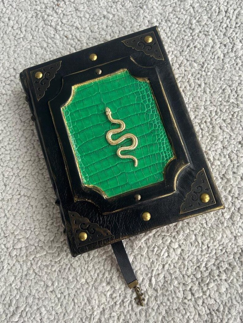 Pode incluir: Um di&aacute;rio encadernado em couro preto com uma capa verde em relevo e um design de cobra dourada. O di&aacute;rio tem um marcador de fita preta e detalhes met&aacute;licos dourados.