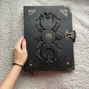 May include: Black leather-bound journal with ornate bronze-colored embellishments. The cover features a triple moon design, stars, and decorative corner pieces. A clasp secures the journal, and a bracelet with black beads is visible.
