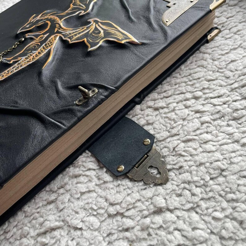 Pu&ograve; includere: Diario in pelle nera con un design a drago dorato. Il diario ha una chiusura a fermaglio e dettagli in metallo color oro. Le pagine sono visibili e hanno un colore legno naturale. Il diario &egrave; su una superficie grigia e morbida.
