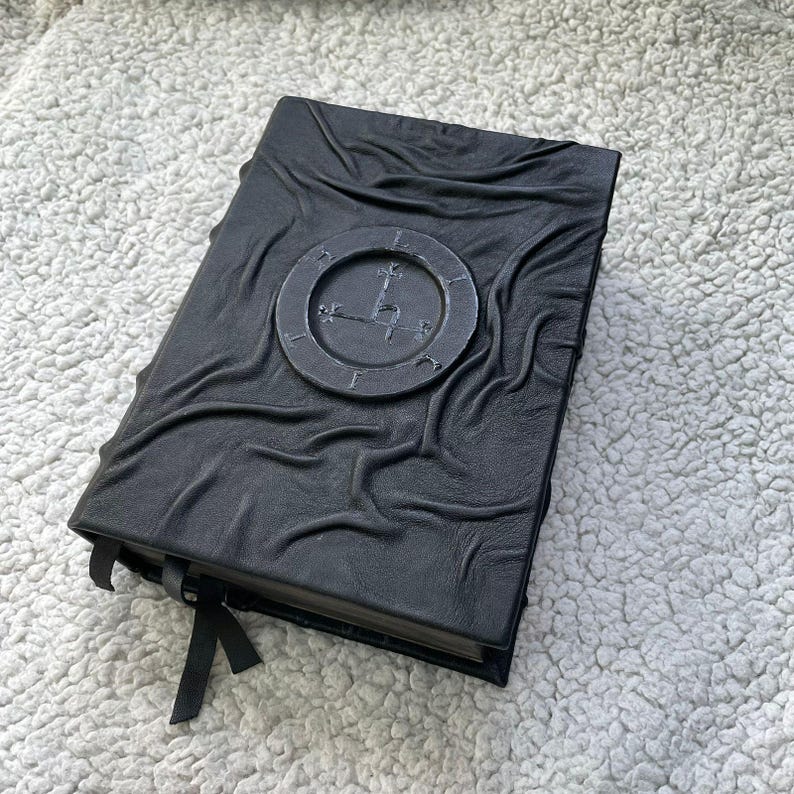 May include: Black leather-bound journal with a textured cover and a circular emblem. The emblem features a stylized clock design with Roman numerals and a cross. A black ribbon bookmark is attached.