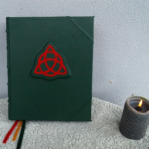 May include: A dark green leather-bound journal with a red embossed triquetra symbol on the front cover.