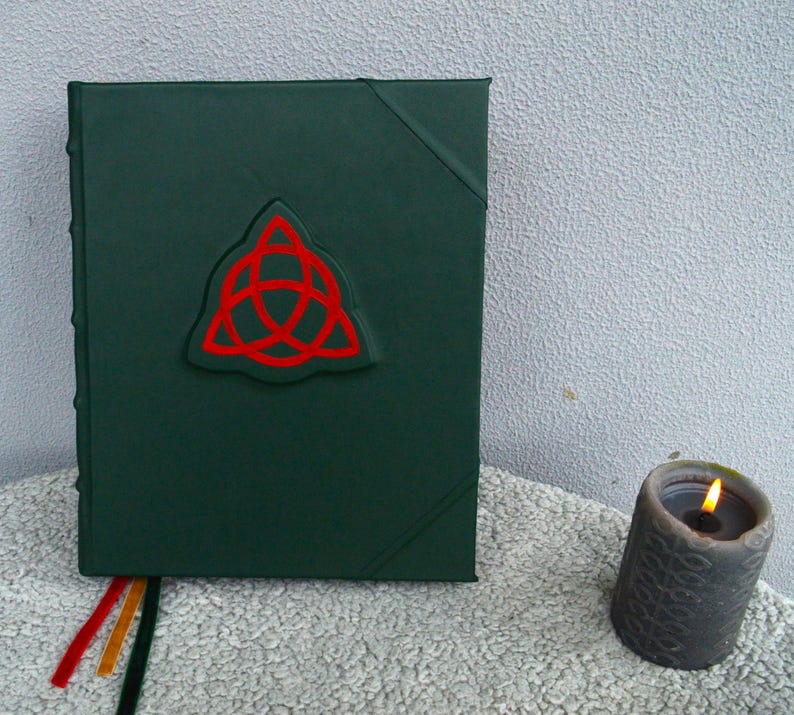 May include: A dark green leather-bound journal with a red embossed triquetra symbol on the front cover.