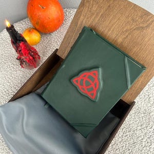 May include: A dark green leather-bound journal with a red triquetra symbol on the cover. The journal is in a wooden box with a gray velvet lining.