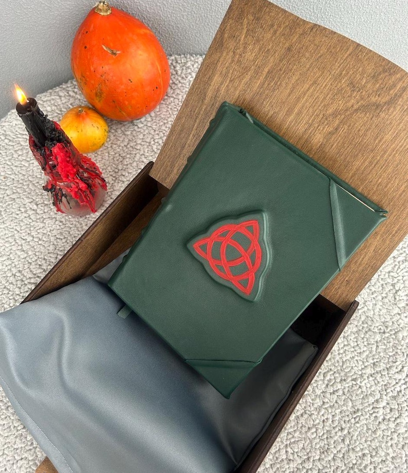 May include: A dark green leather-bound journal with a red triquetra symbol on the cover. The journal is in a wooden box with a gray velvet lining.