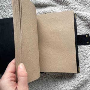 May include: A black journal with a flap closure and brass corner protectors. The pages are a light brown color, and the journal is open, revealing the blank pages. The journal is on a textured, light gray surface.