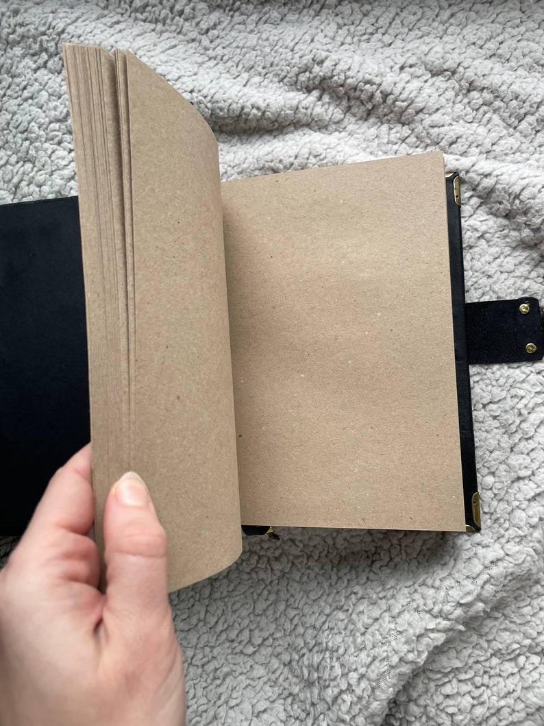May include: A black journal with a flap closure and brass corner protectors. The pages are a light brown color, and the journal is open, revealing the blank pages. The journal is on a textured, light gray surface.