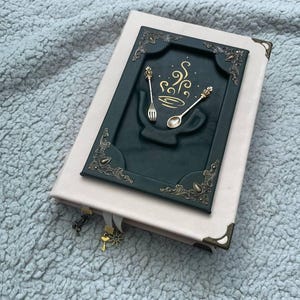 Puede incluir: Un libro rosa claro con una incrustación rectangular verde oscuro en la portada. La incrustación presenta una forma de taza verde oscuro en relieve, un tenedor dorado, una cuchara dorada y líneas de vapor doradas en espiral.