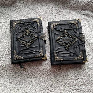May include: Two black leather-bound journals with ornate metal corner accents and clasps. The covers feature embossed designs and the word "Grimoire" in a decorative font. The journals have a gothic or fantasy theme.