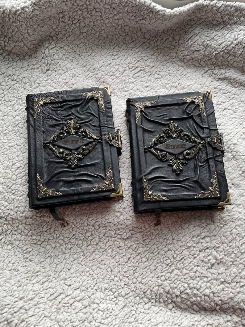 May include: Two black leather-bound journals with ornate metal corner accents and clasps. The covers feature embossed designs and the word "Grimoire" in a decorative font. The journals have a gothic or fantasy theme.