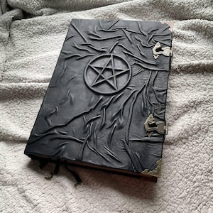 Könnte beinhalten: Schwarzes, ledergebundenes Notizbuch mit einem erhabenen Pentagramm-Design. Der Einband hat ein strukturiertes, faltiges Aussehen und ist mit Metallverschlüssen und Eckschützern gesichert. Ein Leseband ist sichtbar.