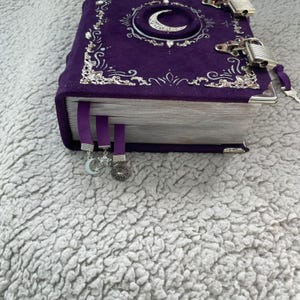 May include: A purple velvet journal with silver detailing, a crescent moon motif, and two padlocks. The journal features three purple ribbon bookmarks with celestial charms, including a moon, star, and sun.