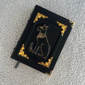 Puede incluir: Diario de cuero negro con detalles dorados y una imagen en relieve de un gato negro con un collar dorado.