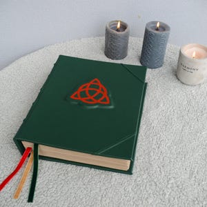 May include: A dark green leather-bound journal with a red triquetra symbol on the front cover. The journal has three tan ribbon bookmarks and is sitting on a white fluffy surface. Three candles are in the background.