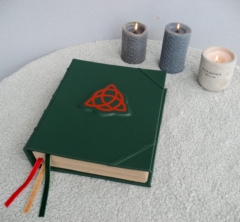 May include: A dark green leather-bound journal with a red triquetra symbol on the front cover. The journal has three tan ribbon bookmarks and is sitting on a white fluffy surface. Three candles are in the background.