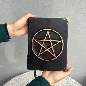 May include: Black leather journal with a gold pentagram design on the front cover. The journal has brass corner accents and a leather strap closure.