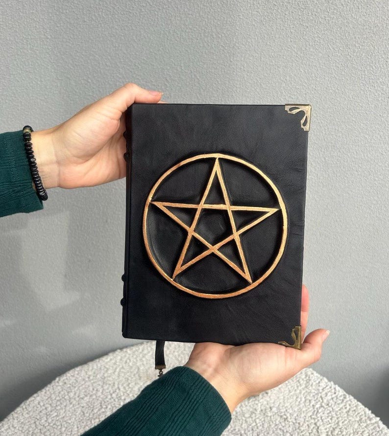 May include: Black leather journal with a gold pentagram design on the front cover. The journal has brass corner accents and a leather strap closure.