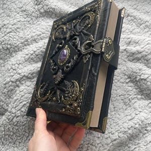 May include: Black leather journal with ornate brass accents and a purple gemstone. The journal features a clasp closure and corner protectors. The cover is embossed with intricate designs, creating a vintage and gothic aesthetic.