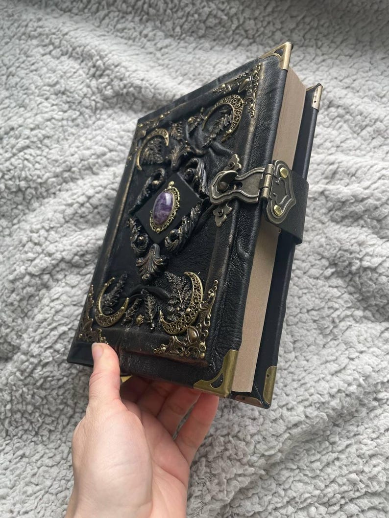 May include: Black leather journal with ornate brass accents and a purple gemstone. The journal features a clasp closure and corner protectors. The cover is embossed with intricate designs, creating a vintage and gothic aesthetic.