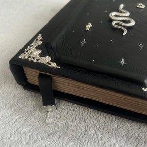 Op de afbeelding: Een zwart lederen gebonden dagboek met zilveren en gouden accenten. De omslag heeft een zilveren slangontwerp en witte sterren. Het dagboek heeft een zwart lint bladwijzer met een kristallen hanger.