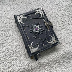May include: A black, embossed journal with silver accents, including crescent moons and decorative corners. The journal features a central, ornate design with a purple gemstone and a metal clasp closure. A black ribbon bookmark is visible.