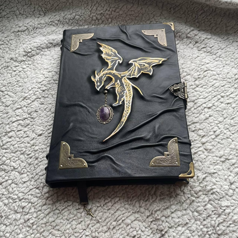 Pu&ograve; includere: Diario rilegato in pelle nera con un emblema di drago oro e nero e un ciondolo con pietra preziosa viola. Il diario presenta angoli decorativi e una chiusura a fermaglio. Un segnalibro con un ciondolo a stella &egrave; appeso in basso.