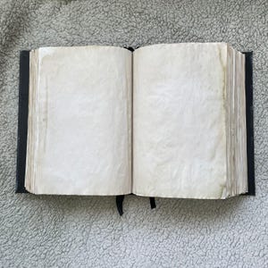 May include: An open, antique-style book with aged, off-white pages. The book has a black cover and a black ribbon bookmark. The pages show signs of age, with slight discoloration and a textured appearance, suggesting it's a vintage or handcrafted item.