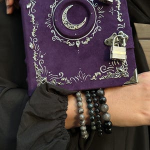 May include: A purple journal with silver embellishments and a crescent moon design. The journal has two silver padlocks and is held with multiple beaded bracelets. The journal's cover is decorated with ornate silver filigree.
