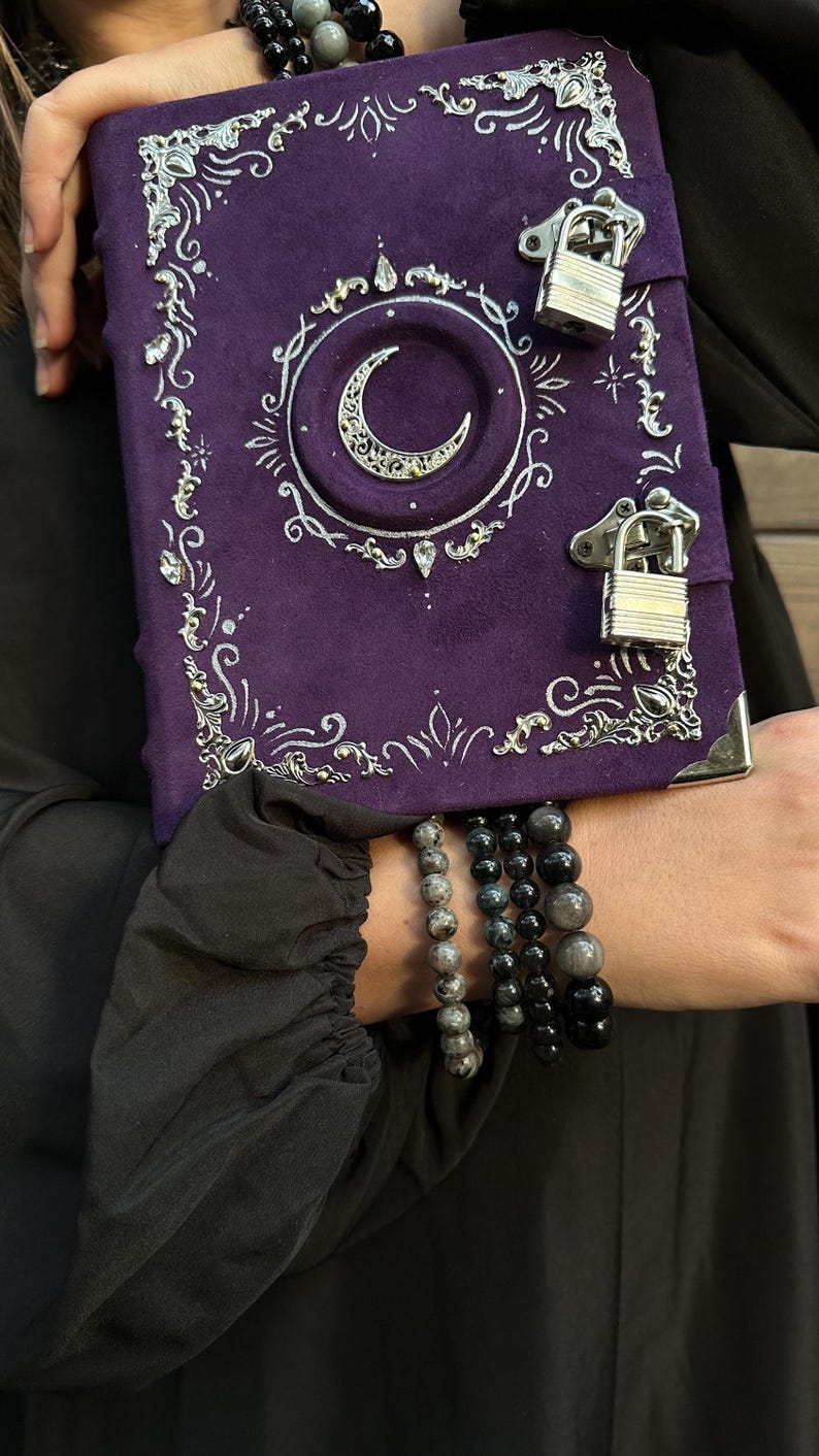 May include: A purple journal with silver embellishments and a crescent moon design. The journal has two silver padlocks and is held with multiple beaded bracelets. The journal's cover is decorated with ornate silver filigree.