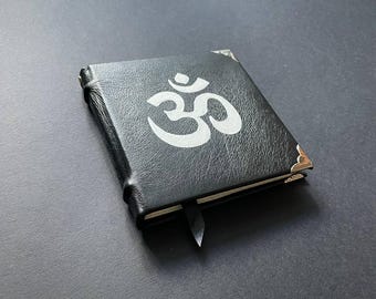 Könnte beinhalten: Ein in schwarzes Leder gebundenes Notizbuch mit einem weißen Om-Symbol auf dem Vorderdeckel. Das Notizbuch verfügt über metallische Eckschoner und ein schwarzes Lesebändchen, das an der Unterkante des Buchrückens hervorsteht.