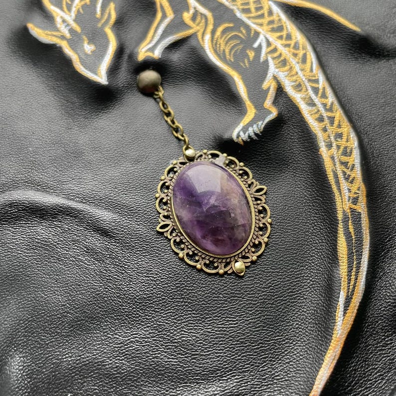 Pu&ograve; includere: Un ciondolo in stile vintage con una grande pietra di ametista ovale incastonata in una cornice in filigrana ornata color bronzo. Il ciondolo &egrave; appeso a una delicata catena, su una superficie in pelle nera con arte del drago dorato e bianco.