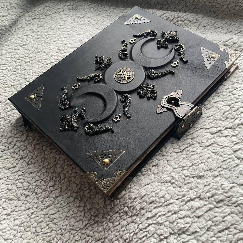 May include: Black leather journal with embossed celestial design featuring moons, stars, and a central circular emblem. The journal has decorative corner accents and a metal clasp closure. A unique, handcrafted item.