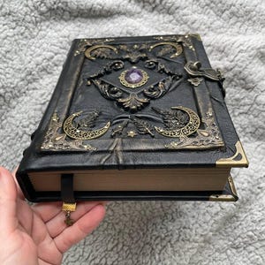 May include: Black leather-bound journal with ornate brass accents, including crescent moons and a central purple gemstone. The journal features a metal clasp closure and a decorative bookmark with a star charm.