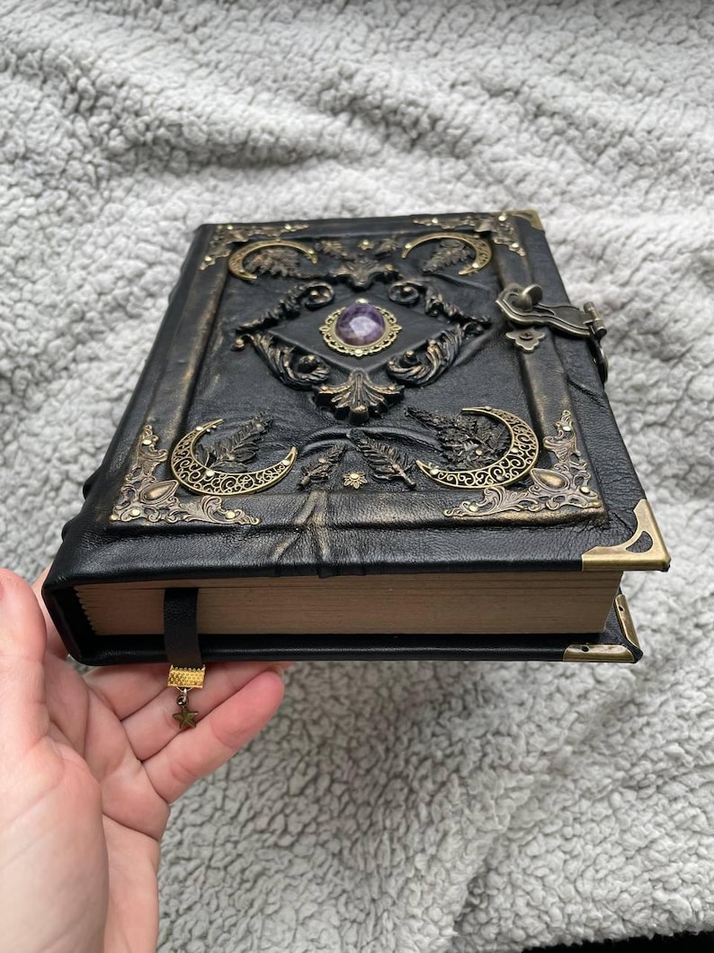 May include: Black leather-bound journal with ornate brass accents, including crescent moons and a central purple gemstone. The journal features a metal clasp closure and a decorative bookmark with a star charm.