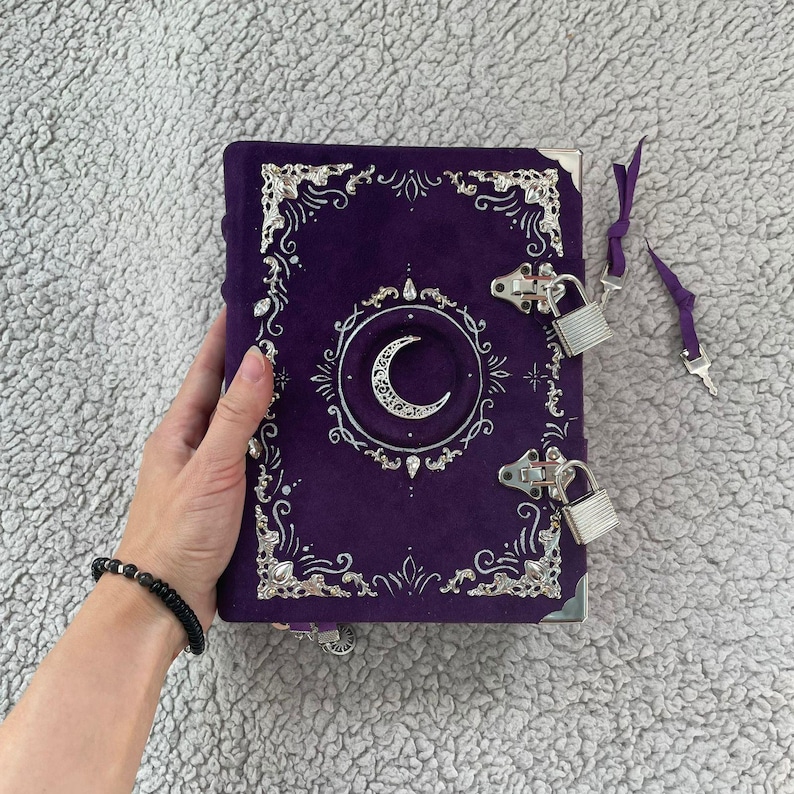May include: A purple journal with a crescent moon design and silver accents. The journal has a velvet-like texture, metal clasps, and a lock. The cover features ornate silver filigree and a decorative border. A purple ribbon is attached.