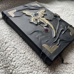 Pu&ograve; includere: Diario rilegato in pelle nera con un ornamento a forma di drago dorato e un ciondolo di ametista. Il diario presenta protezioni angolari decorative e un segnalibro a forma di stella. La copertina ha un design in rilievo strutturato.