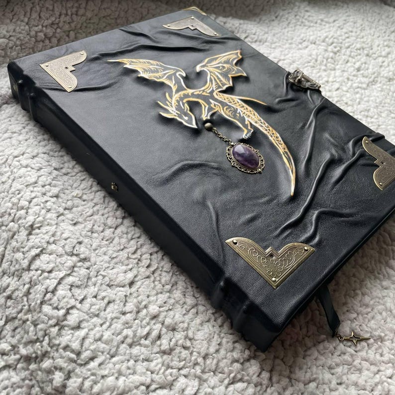 Pu&ograve; includere: Diario rilegato in pelle nera con un ornamento a forma di drago dorato e un ciondolo di ametista. Il diario presenta protezioni angolari decorative e un segnalibro a forma di stella. La copertina ha un design in rilievo strutturato.