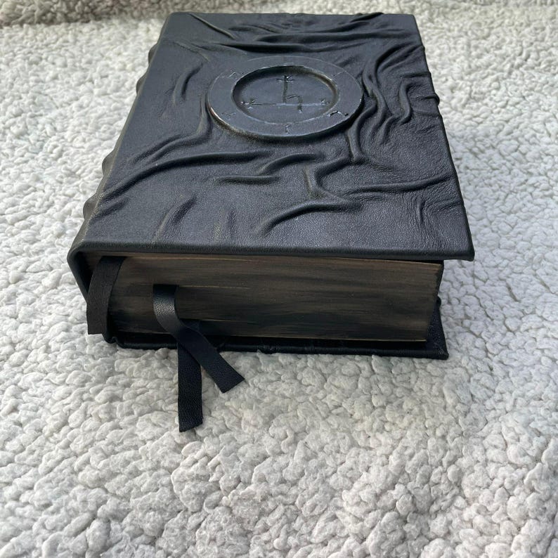 May include: A black leather-bound book with a textured cover and a silver emblem. The book has a black ribbon bookmark and aged-looking pages. The cover has a unique, wrinkled design.