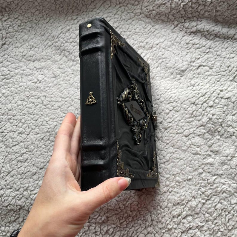 May include: A black, ornate book with gold-colored metal accents. The cover features embossed designs and a decorative clasp. A small, gold-colored emblem is visible on the spine. The book is held by a hand.
