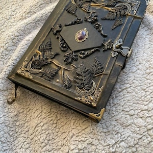 May include: A dark green journal with ornate metal corner accents and a clasp. The cover features embossed fern and leaf designs, with a central diamond-shaped panel holding a purple gemstone. The journal has a decorative metal edge.