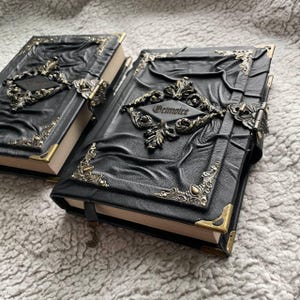 May include: Two black leather-bound books with ornate metal corner accents and clasps. The covers feature embossed designs and a decorative metal plaque that reads "Grimoire". The pages are edged in gold, and a black ribbon bookmark is attached.