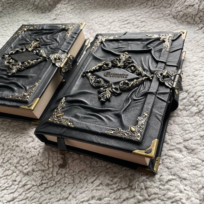 May include: Two black leather-bound books with ornate metal corner accents and clasps. The covers feature embossed designs and a decorative metal plaque that reads "Grimoire". The pages are edged in gold, and a black ribbon bookmark is attached.