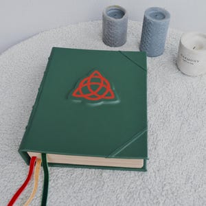 May include: A green leather-bound journal with a red triquetra symbol embossed on the front cover. The journal has three ribbon bookmarks in red, yellow, and green.