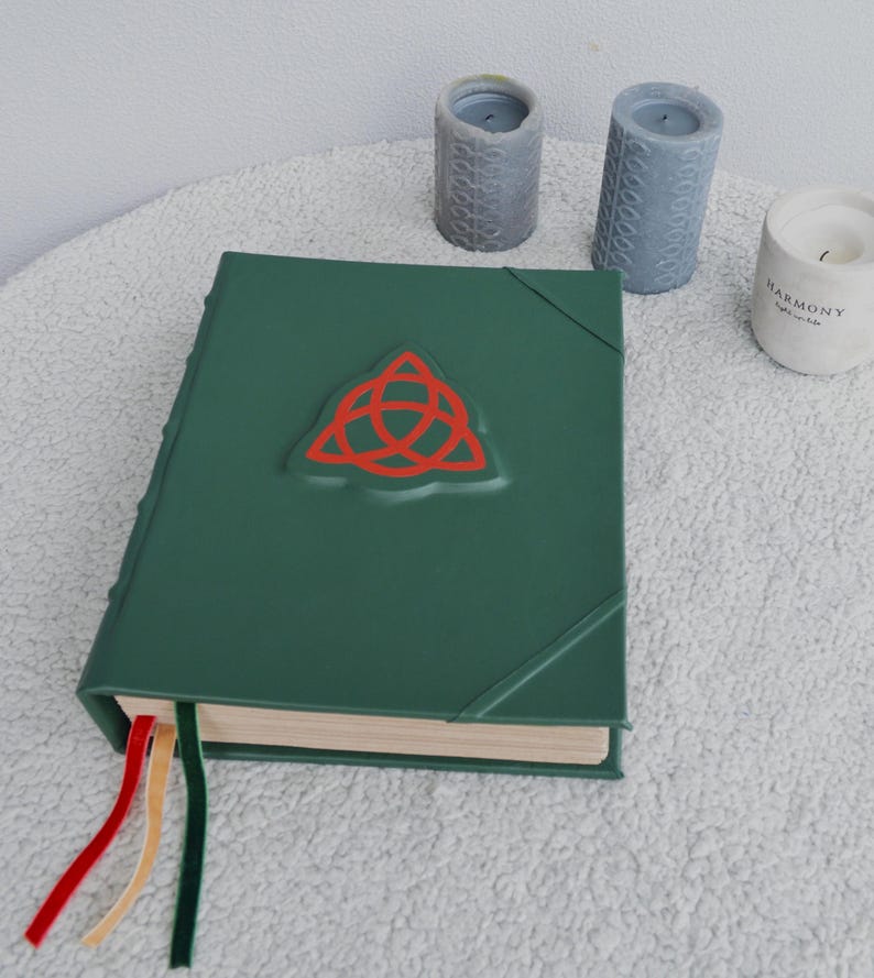 May include: A green leather-bound journal with a red triquetra symbol embossed on the front cover. The journal has three ribbon bookmarks in red, yellow, and green.