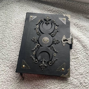May include: Black leather journal with ornate bronze-colored metal accents. The cover features a triple moon design, stars, and decorative flourishes. The journal has a metal clasp closure and corner protectors.