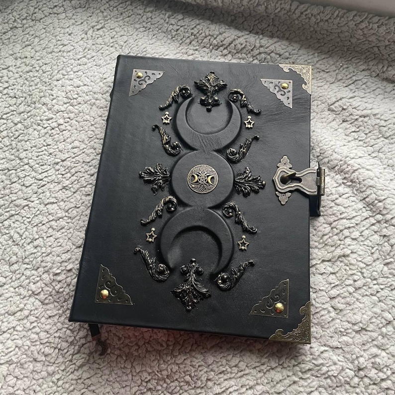May include: Black leather journal with ornate bronze-colored metal accents. The cover features a triple moon design, stars, and decorative flourishes. The journal has a metal clasp closure and corner protectors.
