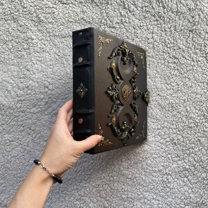 K&ouml;nnte beinhalten: Ein braunes und schwarzes ledergebundenes Buch mit aufwendigen goldfarbenen Metallverzierungen. Das Buch hat einen dekorativen Verschluss und Eckakzente. Der Buchr&uuml;cken ist schwarz mit einem goldfarbenen Blumendesign. Das Buch wird von einer Person gehalten.